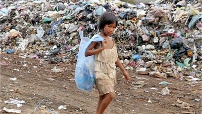 A young Asian girl walks through a rubbish tip with a bag over her shoulder full of things she may be able to use